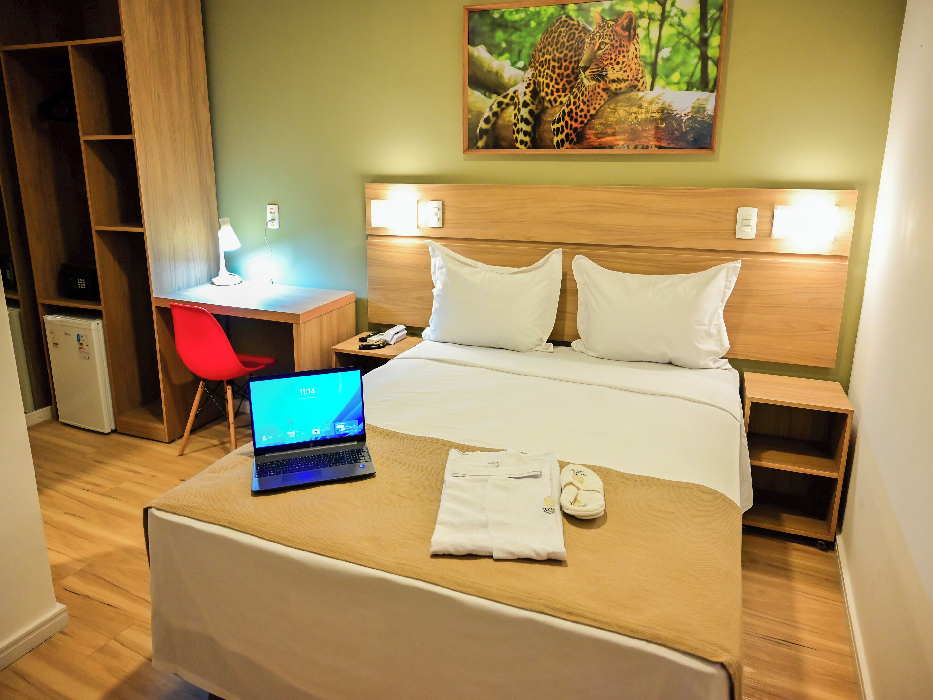 Una habitación de hotel con una cama, un escritorio con una computadora portátil, una silla roja, un refrigerador pequeño y un cuadro en la pared.