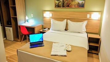 Una habitación de hotel con una cama, un escritorio con una computadora portátil, una silla roja, un refrigerador pequeño y un cuadro en la pared.