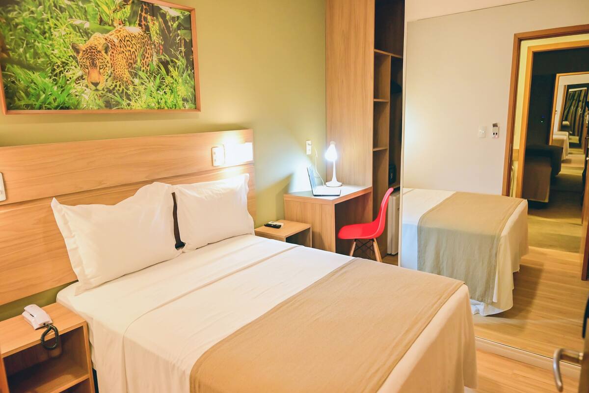 A hotel room with a large bed, a desk with a lamp, a red chair, and a wardrobe.