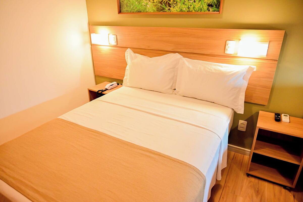 A neatly made bed with white linens, a wooden headboard, bedside tables with lamps, and a small shelf unit.