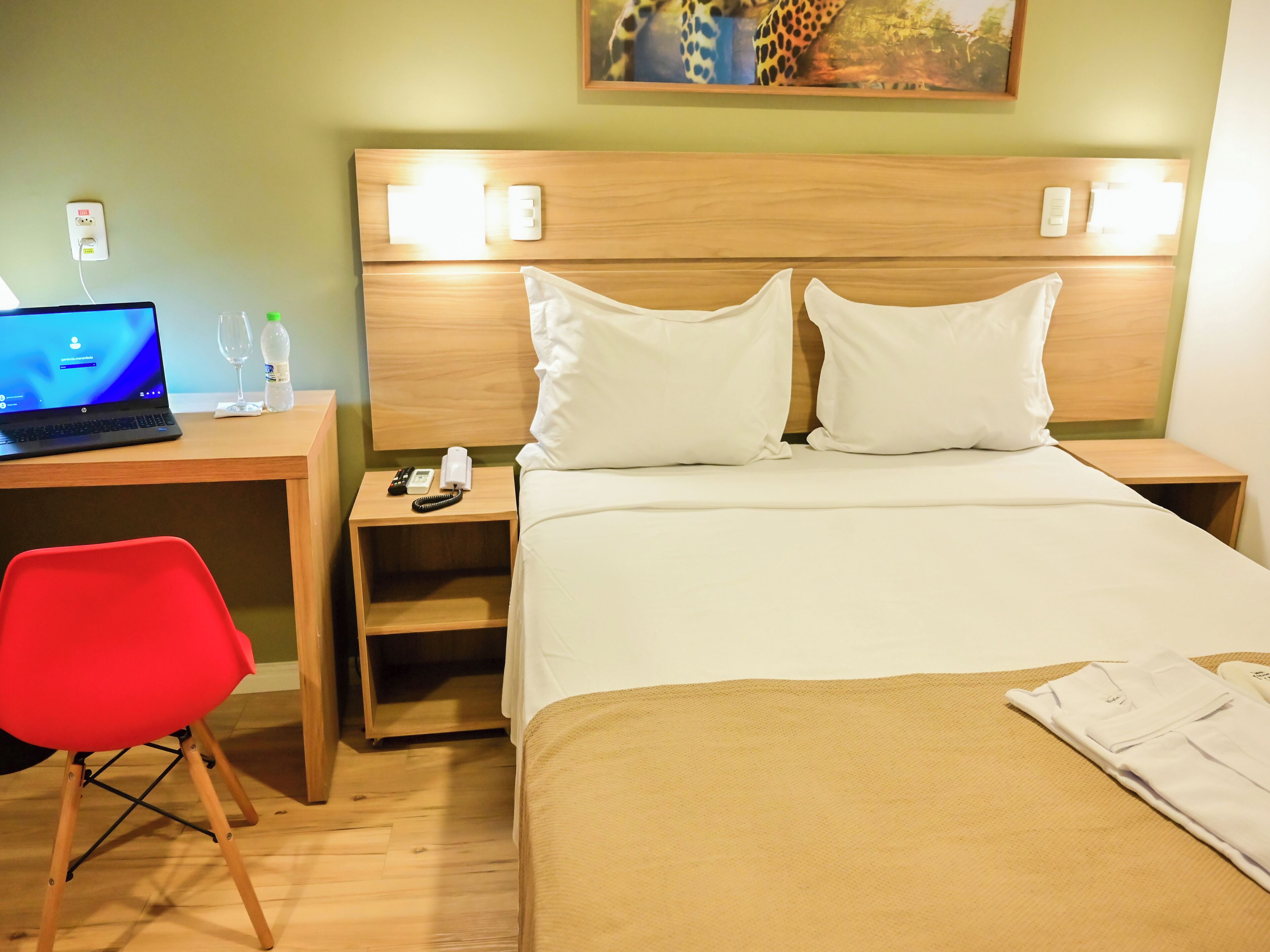 Habitación de hotel con una cama, un escritorio con una computadora portátil, una silla roja y una mesita de noche con un teléfono y una lámpara.