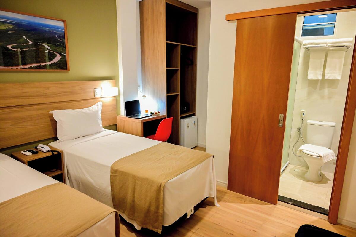 A hotel room with two beds, a desk with a laptop, a red chair, a wardrobe, and a shower area.