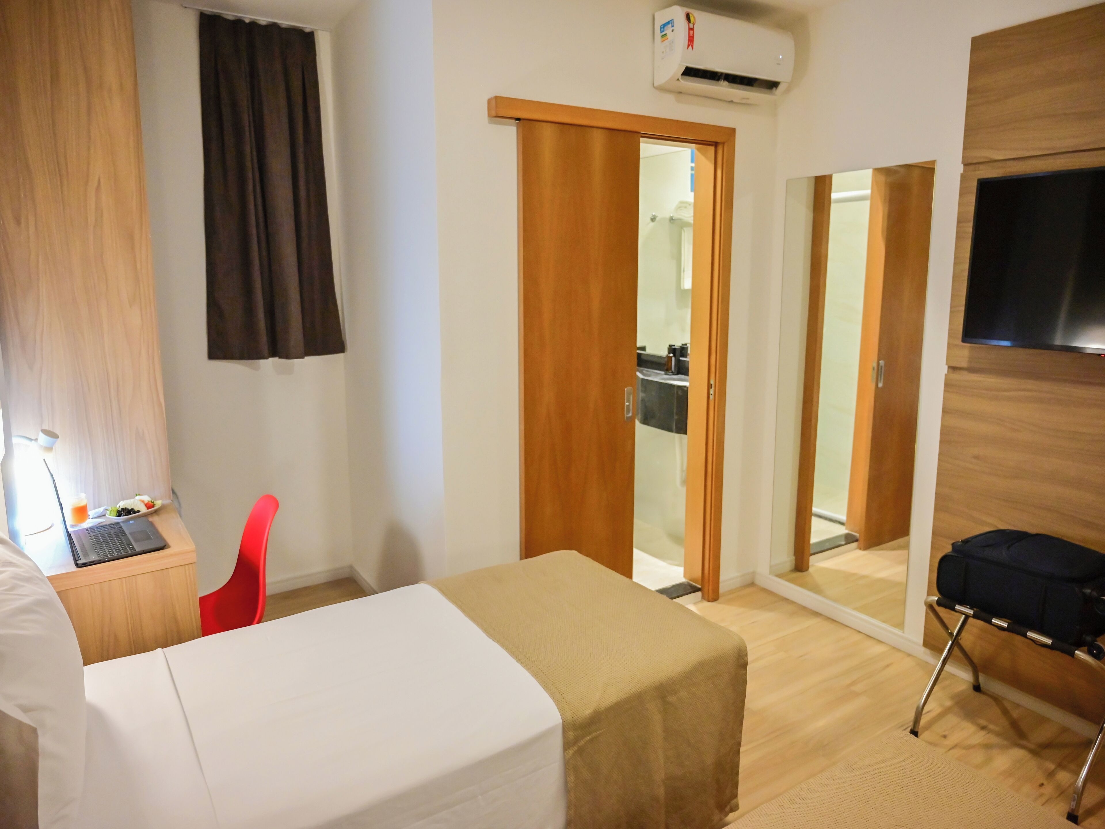 A hotel room with a bed, a desk with a laptop, a red chair, a TV mounted on the wall, and a sliding door leading to a bathroom.