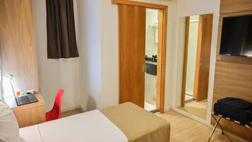 A hotel room with a bed, a desk with a laptop, a red chair, a TV mounted on the wall, and a sliding door leading to a bathroom.