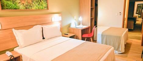 A hotel room with a large bed, a desk with a lamp, a red chair, and a wardrobe.