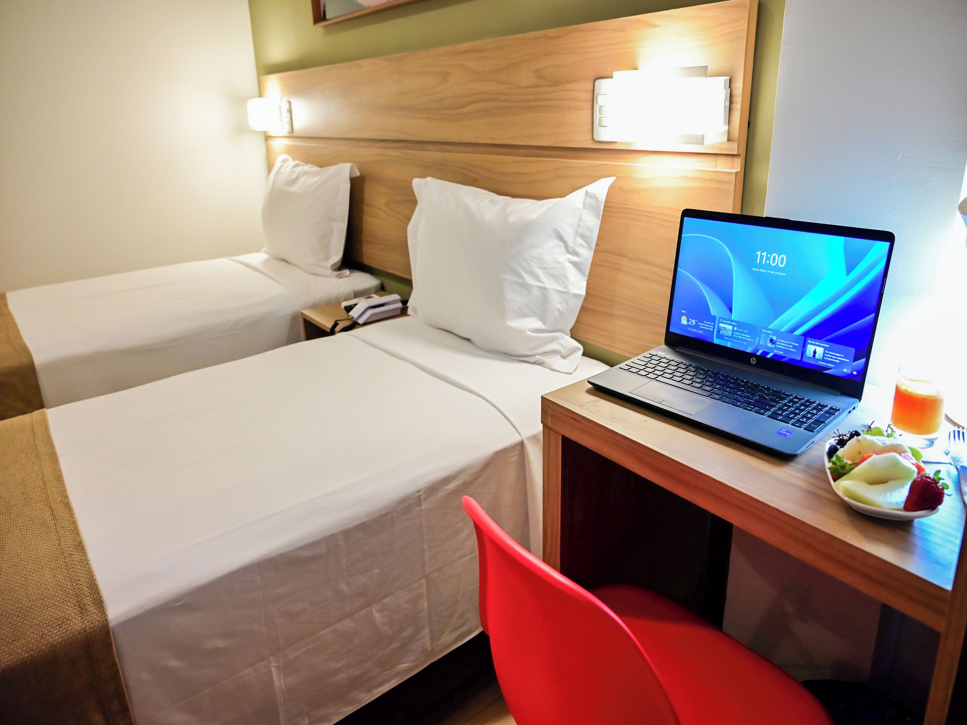 A hotel room with two beds, a laptop on a desk, and a fruit plate.