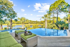 An infinity pool with a wooden deck, wicker lounge chairs, and a view of a lake surrounded by trees.