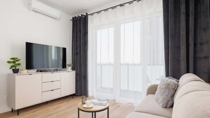 Ein modernes Wohnzimmer mit einem Flachbildfernseher auf einer weißen Kommode, einem beige farbenen Sofa, einem kleinen runden Couchtisch und transparenten Vorhängen.