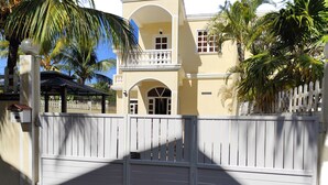 A gated entrance with a white gate, a yellow building with a balcony, and palm trees.