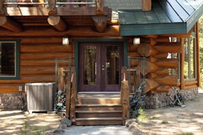 A log cabin with a wooden staircase leading to a front door, a stone foundation, and a metal air conditioning unit. - Riverfront Log Cabin with Firepit in La Pine (La Pine)