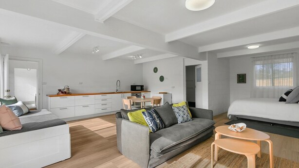 Une pièce moderne et bien éclairée, avec un canapé gris, une petite table ronde et un lit. On aperçoit à l’arrière-plan un coin cuisine doté de meubles blancs et d’un four à micro-ondes.