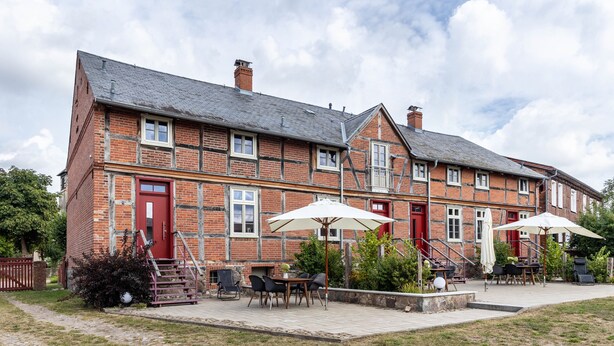 A brick building with a red door, outdoor seating area with tables and chairs, and a staircase leading to the entrance. - Apartment '1' with Private Terrace, Shared Garden and Wi-Fi (Groß Pankow)