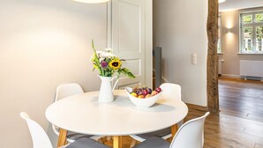 Ein Essbereich mit rundem Tisch, weißen Stühlen, einer Blumenvase und einer Obstschale.