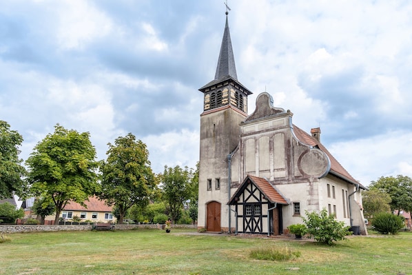 A church with a tall steeple, a small adjacent building, and a grassy area with trees.