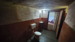 Une petite salle de bain étroite et peu éclairée, équipée de toilettes, d’un lavabo et d’une poubelle verte.