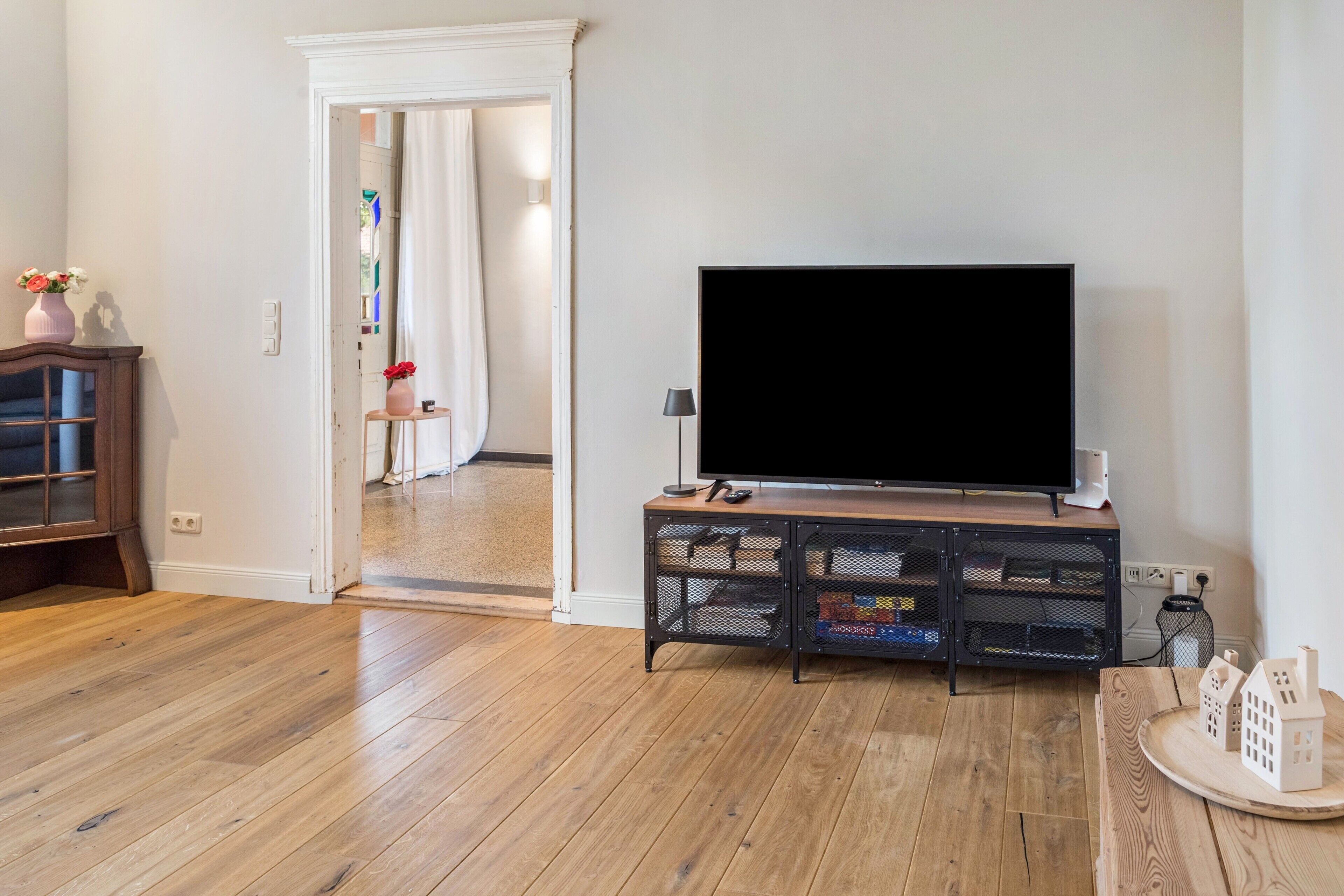 A living room with a flat-screen TV on a wooden stand, a small table with decorative houses, and a doorway leading to another room.