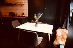 Un coin repas avec une table blanche, deux chaises, un vase de fleurs et, au fond, une cuisine.