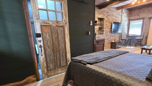 Una habitación rústica con una cama grande, una puerta de madera, un muro de ladrillo, una pequeña cocina y un televisor montado en la pared.