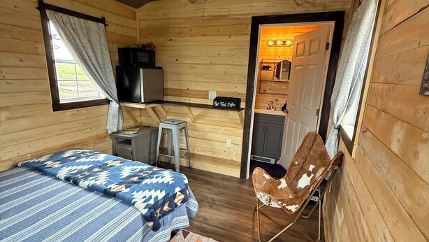 A cozy cabin-style room with a bed, a chair, a small desk, and a kitchenette. - Chief Lone Walker #6 (East Glacier Park)