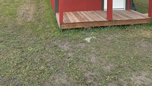 A small red wooden structure with a white door and windows, situated in a grassy field with mountains in the background. - Chief Lone Walker #6 (East Glacier Park)
