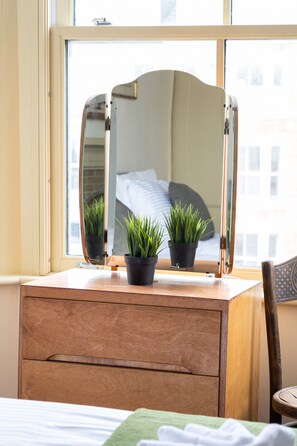 Een houten dressoir met een spiegel, twee planten in potten, en een bed met een groene deken.
