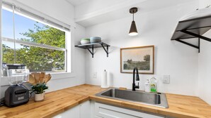 A modern kitchen with a wooden countertop, white cabinets, a stainless steel sink, a blender, a potted plant, and a hanging pendant light.
