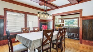 Un comedor con una mesa y sillas de madera, una araña y ventanas amplias con persianas.