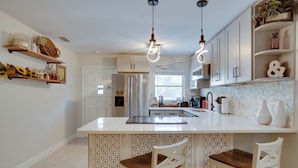 Una cocina moderna con armarios blancos, una isla central con dos sillas de madera, iluminación colgante y objetos decorativos en estantes.