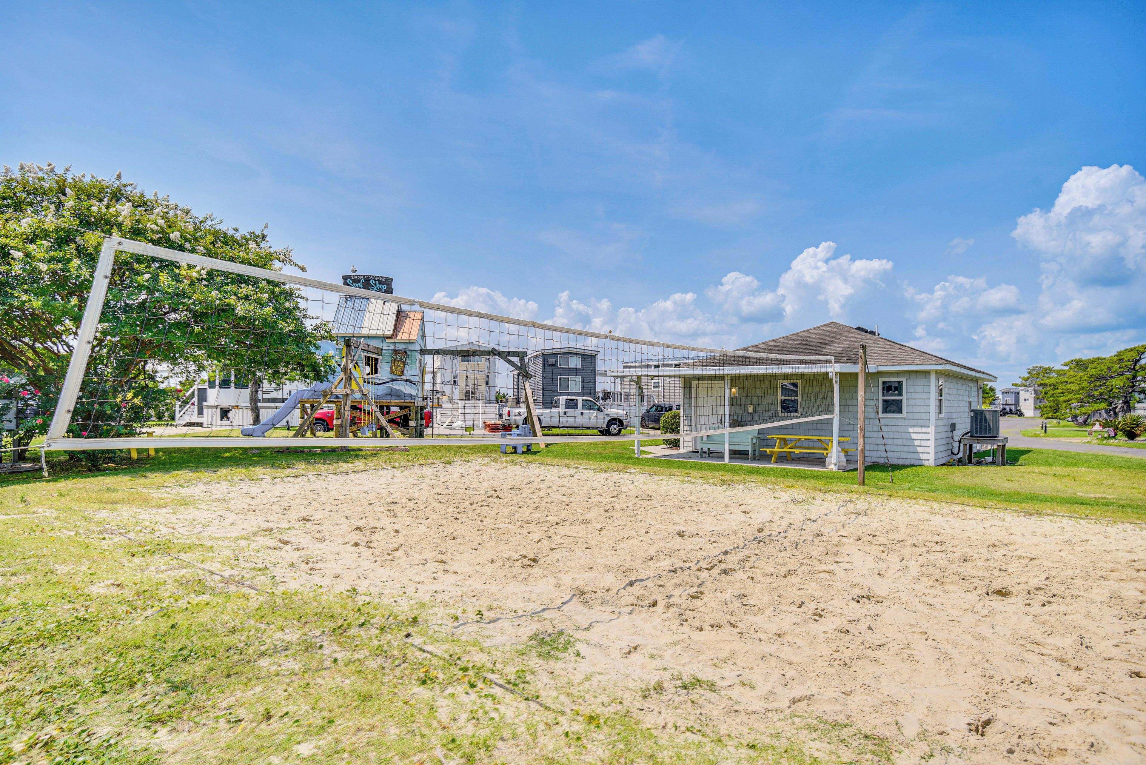 A sandy volleyball court with a net, a small building with a porch, and a tree in the background.