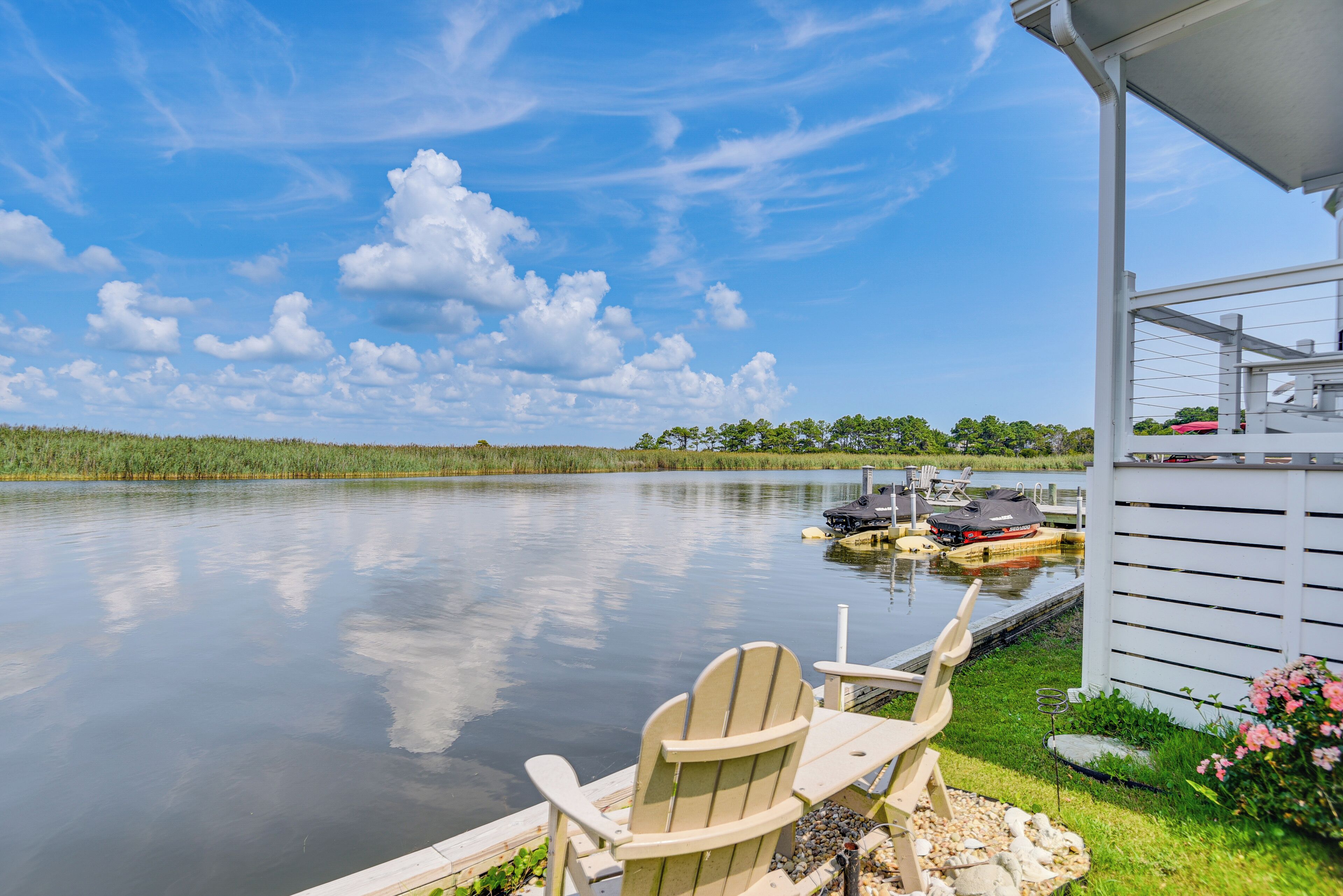 A lakeside deck with Adirondack chairs, a dock with boats, and a small building with a sloped roof.