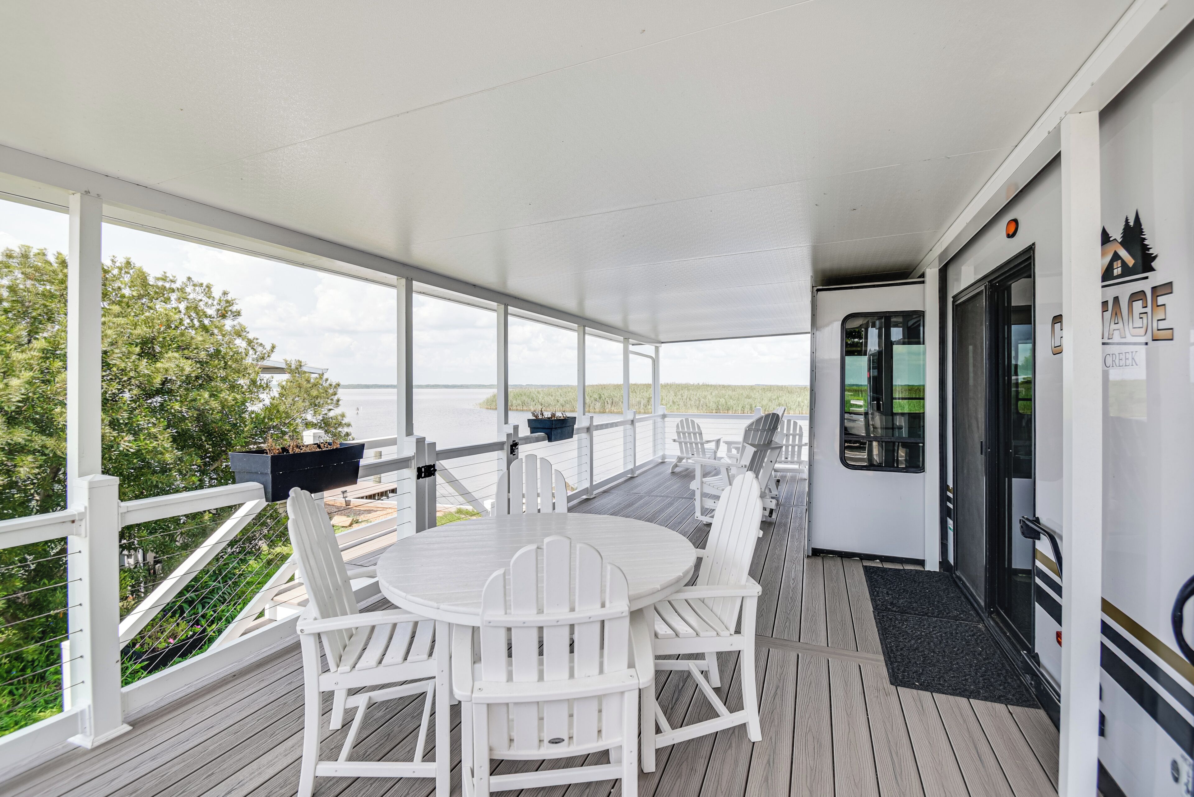 Une terrasse extérieure couverte, dotée d’une table ronde et de chaises Adirondack blanches, qui offre une vue sur une pelouse et un plan d’eau.