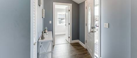 Un pasillo con suelo de madera, una consola blanca con un florero y una puerta que da a otra habitación.