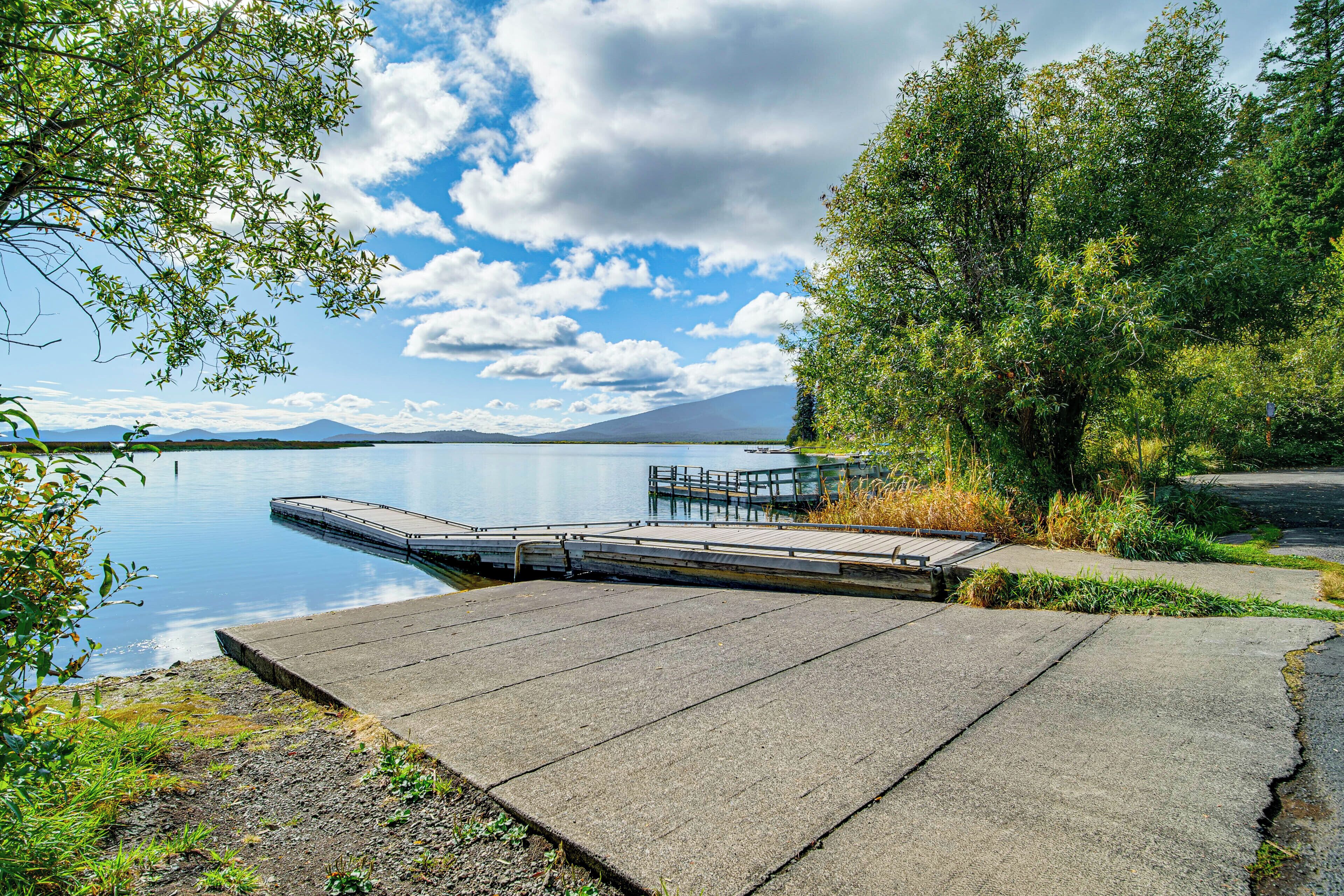 A concrete dock extending into calm water, surrounded by greenery and a clear sky.