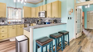 A kitchen with yellow cabinets, a stainless steel trash can, and a bar area with stools. - Walk to the Ocean: Family Retreat in Crystal Beach (Port Bolivar)