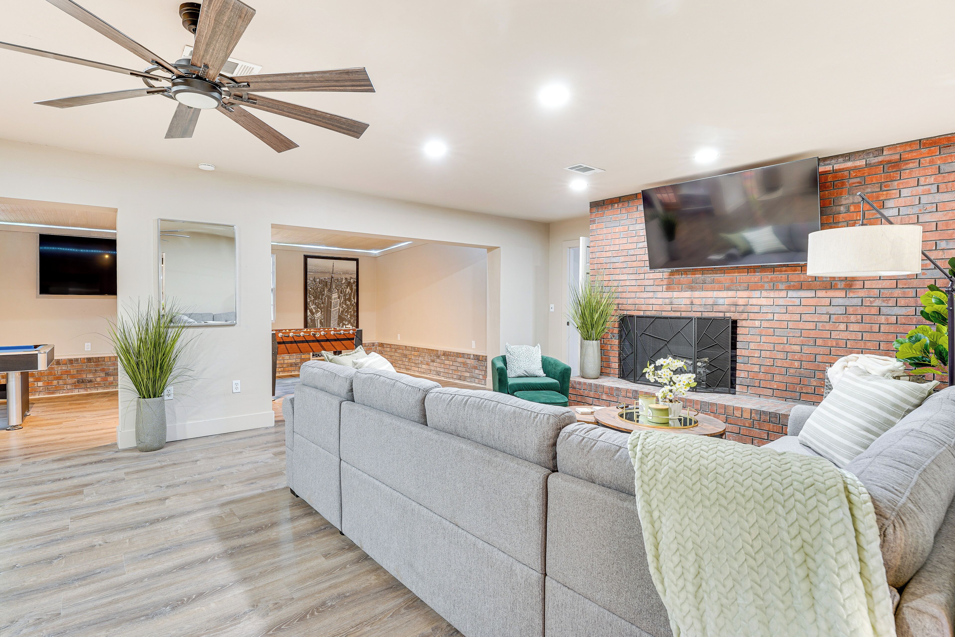 A modern living room with a sectional sofa, a flat-screen TV mounted on a brick wall, a ceiling fan, and a fireplace.