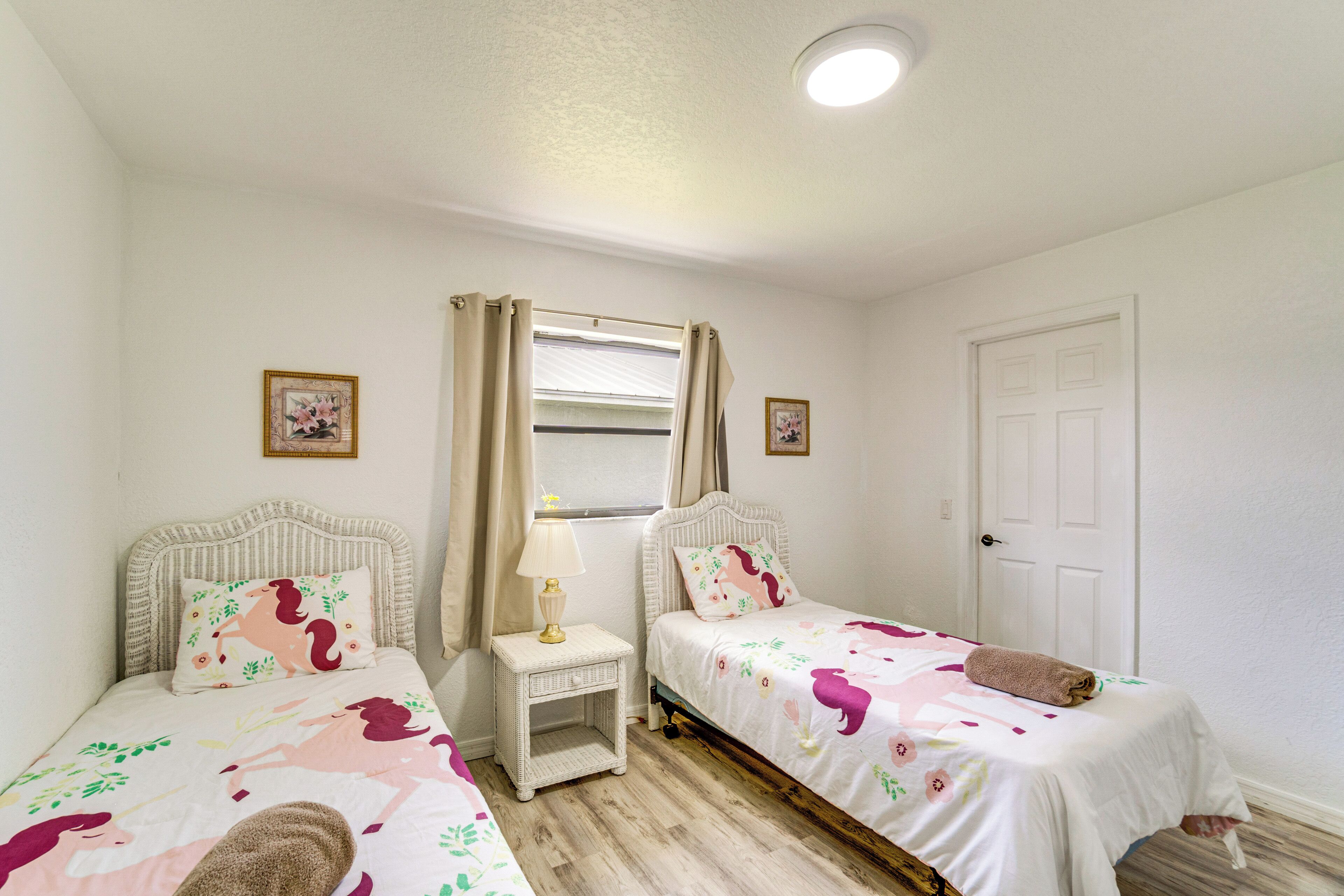 A bedroom with two beds, a window with curtains, a bedside table with a lamp, and a door.