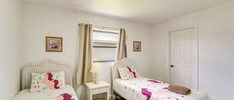 A bedroom with two beds, a window with curtains, a bedside table with a lamp, and a door.
