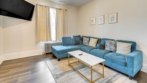 A living room with a blue sectional sofa, a white coffee table, a flat-screen TV mounted on the wall, and a radiator under a window with beige curtains. - 1 Block to Stroudsburg Main St: Chic Townhome (Stroudsburg)