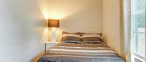 A bedroom with a striped bedspread, a bedside lamp, and a window with blinds.