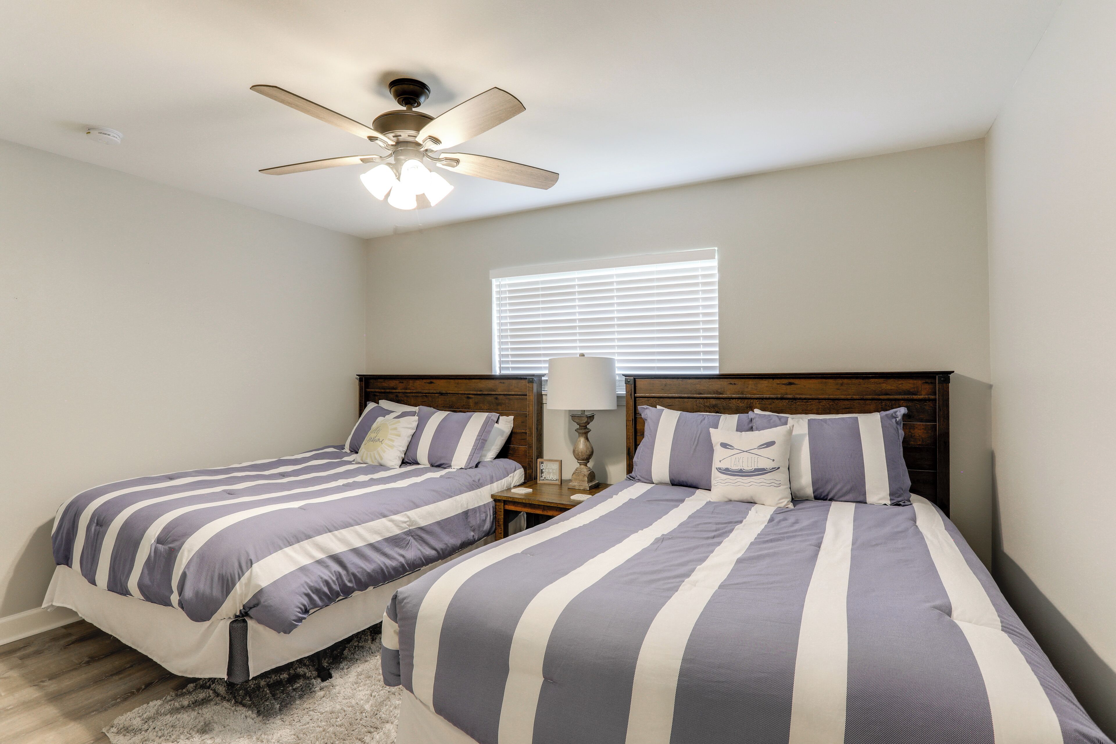A bedroom with two beds, a ceiling fan, a window with blinds, and a nightstand with a lamp.