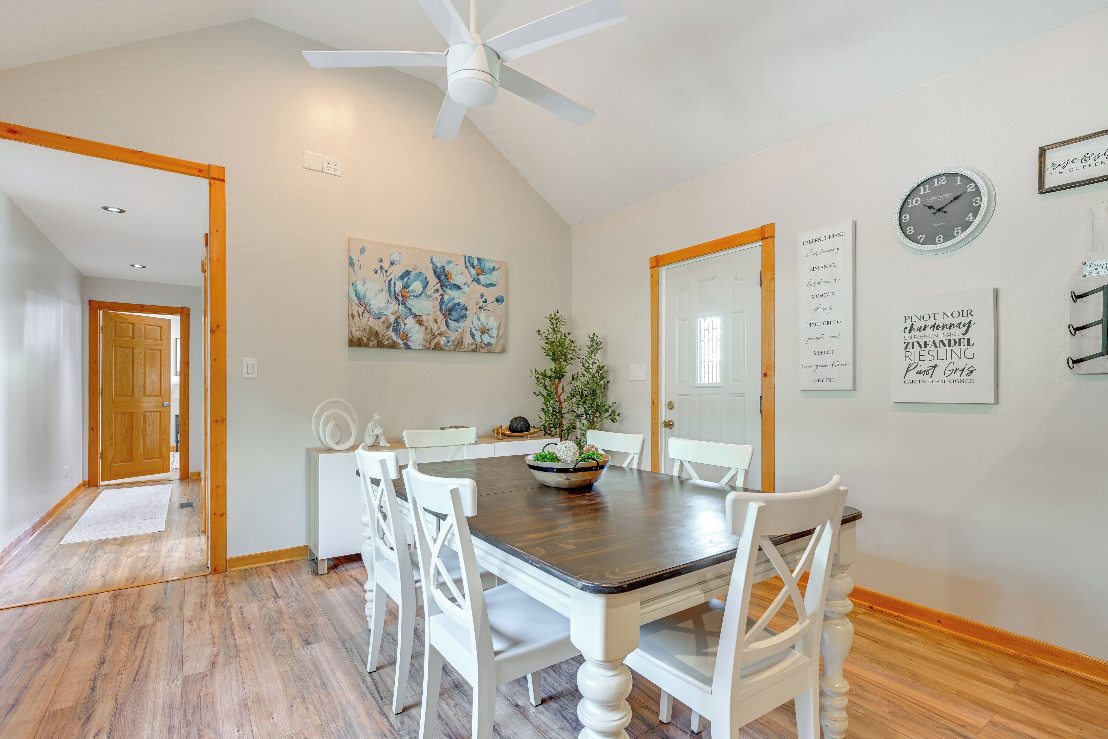 A dining area with a wooden table and white chairs, a ceiling fan, and a painting on the wall.