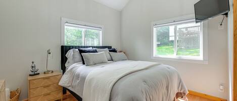 A bedroom with a bed, a nightstand, a TV, and two windows.