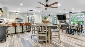 A modern kitchen with a central island, bar stools, and a dining area with chairs and a TV.