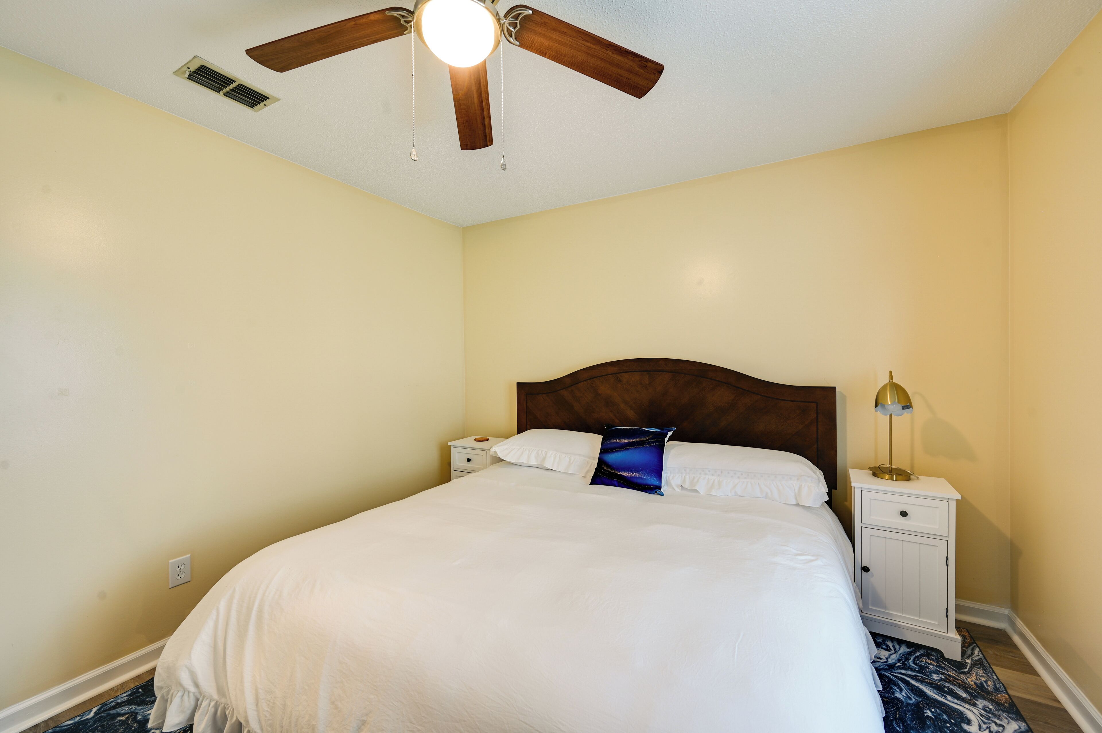 A bedroom with a wooden headboard, a bed with white bedding, a nightstand with a lamp, and a ceiling fan.