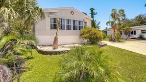 A single-story mobile home with a front porch, surrounded by a well-maintained lawn and various tropical plants. - Tee Off & Soak Up Sun! Airy Englewood Gem w/ Yard (Englewood)