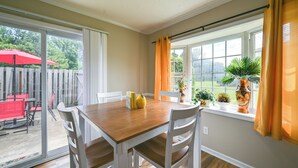 Un comedor con una mesa de madera y sillas blancas, una puerta corrediza de vidrio que da a un patio al aire libre con una sombrilla roja, y una ventana con cortinas naranjas.