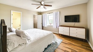 Un dormitorio con una cama, ventilador de techo, cómoda, televisor y ventana con cortinas.