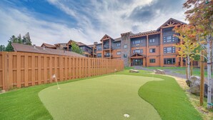 Un parcours de golf avec une clôture en bois, un green d’entraînement et un bâtiment à la façade en pierre en arrière-plan.