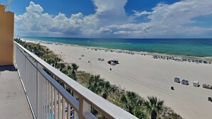 Une vue sur la plage depuis un balcon, avec un ciel dégagé et une plage de sable où l’on aperçoit des parasols et des chaises longues.