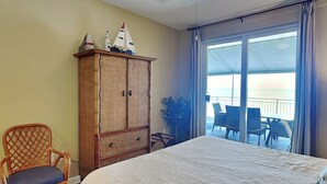 Une chambre avec un lit, une chaise en osier, une armoire et une vue sur l’océan par une porte coulissante.